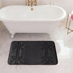 A 20" x 32" Quilted Memory Foam Bath Mat in black, featuring embossed bow and ribbon patterns, is placed on a white marble floor in front of a clawfoot tub with gold fixtures. A small table holding a pink and white item sits nearby.