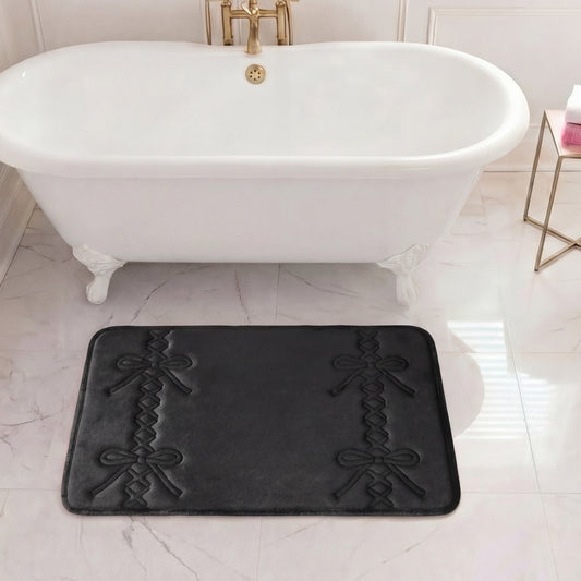 A 20" x 32" Quilted Memory Foam Bath Mat in black, featuring embossed bow and ribbon patterns, is placed on a white marble floor in front of a clawfoot tub with gold fixtures. A small table holding a pink and white item sits nearby.
