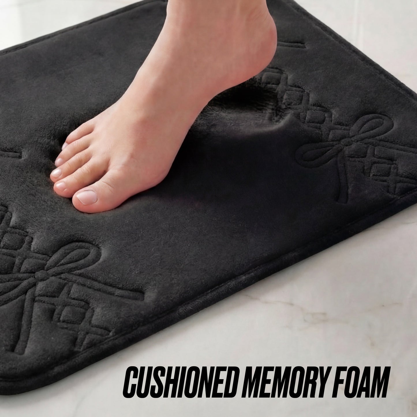 A bare foot steps onto the Quilted Memory Foam Bath Mat, 20" x 32", leaving an impression on its cushioned surface with bow patterns. The words CUSHIONED MEMORY FOAM appear at the bottom, emphasizing its stylish comfort.
