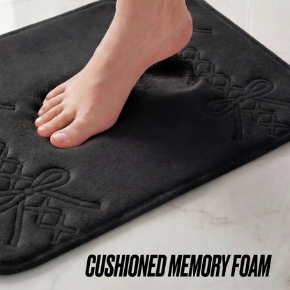 A bare foot steps onto the Quilted Memory Foam Bath Mat, 20" x 32", leaving an impression on its cushioned surface with bow patterns. The words CUSHIONED MEMORY FOAM appear at the bottom, emphasizing its stylish comfort.