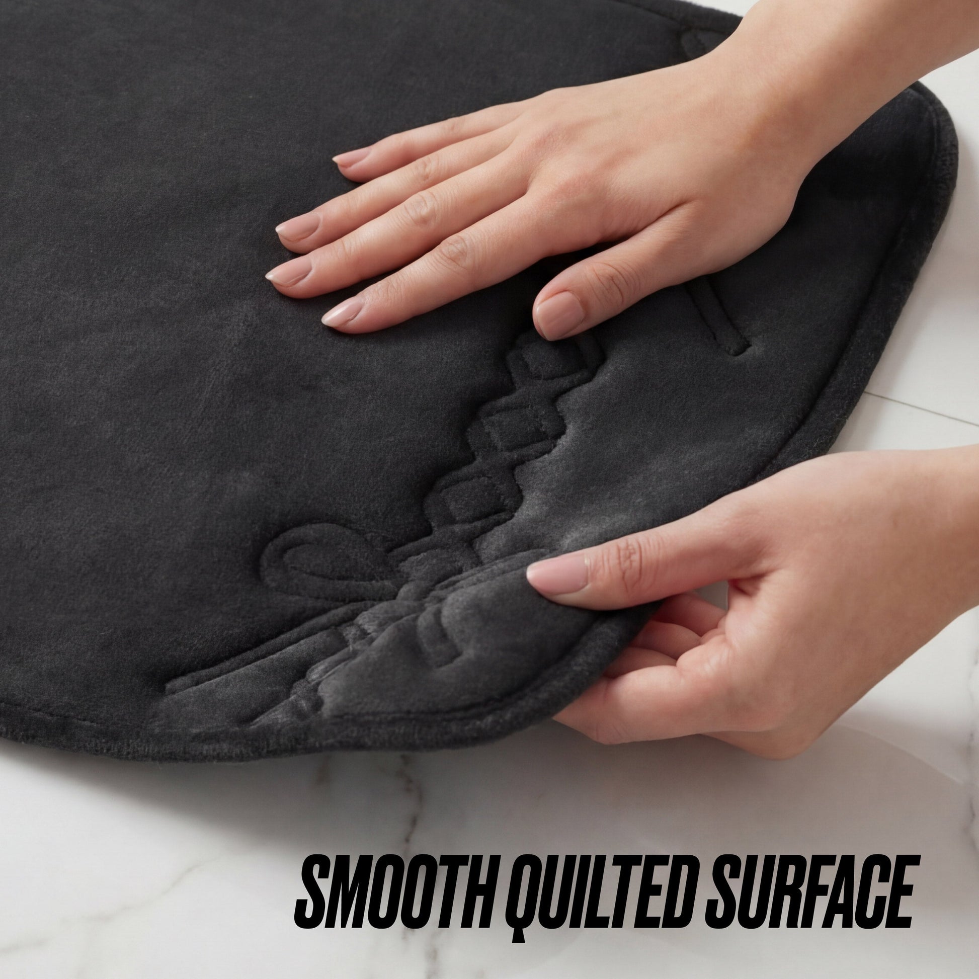 A person's hands touch the corner of the Quilted Memory Foam Bath Mat, 20" x 32", in dark gray on a white surface. The mat features visible stitching and the words SMOOTH QUILTED SURFACE at the bottom.