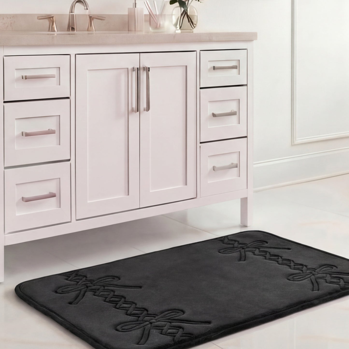 A Quilted Memory Foam Bath Mat (20" x 32"), black with a decorative pattern, is placed on a white tiled floor in front of a white bathroom vanity with chrome handles and a beige countertop.