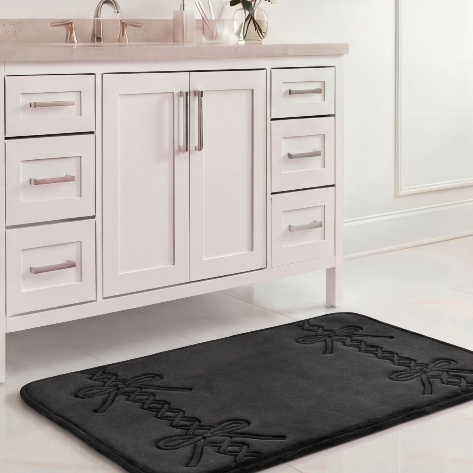 A Quilted Memory Foam Bath Mat (20" x 32"), black with a decorative pattern, is placed on a white tiled floor in front of a white bathroom vanity with chrome handles and a beige countertop.