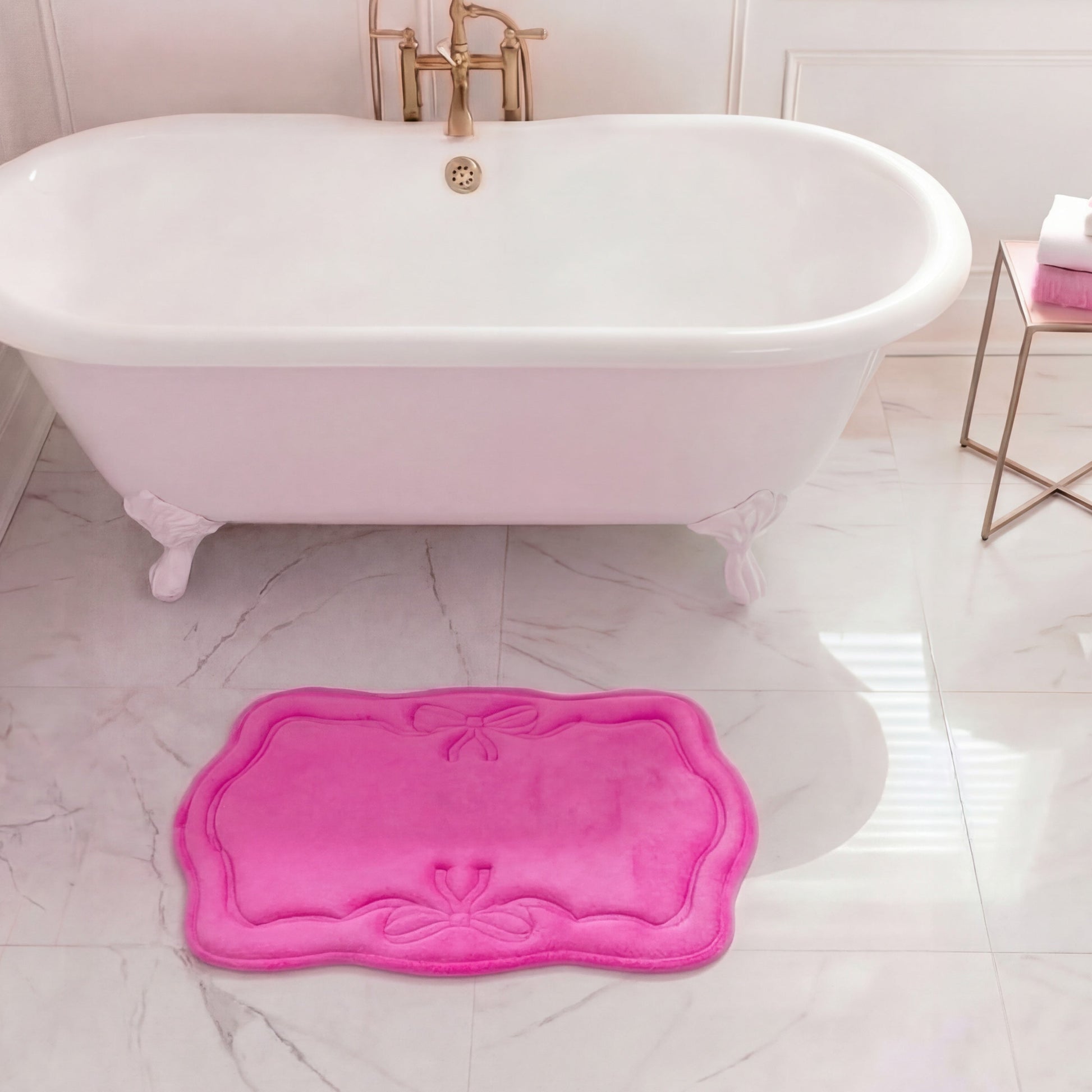 A white clawfoot tub with gold fixtures stands on marble, accented by a bright pink Quilted Memory Foam Bath Mat (17" x 24") in front. To the right, a small table displays folded white and pink towels.