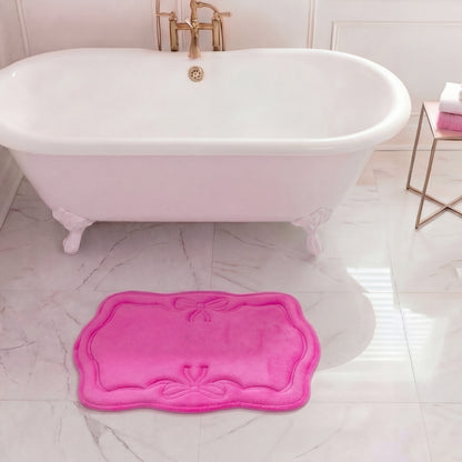 A white clawfoot tub with gold fixtures stands on marble, accented by a bright pink Quilted Memory Foam Bath Mat (17" x 24") in front. To the right, a small table displays folded white and pink towels.