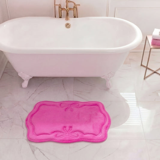A white clawfoot tub with gold fixtures stands on marble, accented by a bright pink Quilted Memory Foam Bath Mat (17" x 24") in front. To the right, a small table displays folded white and pink towels.