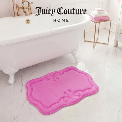 A luxurious white bathtub and gold faucet gleam in a bright bathroom. The Quilted Memory Foam Bath Mat, 17" x 24", by Juicy Couture adds a pink touch on the marble floor. Folded towels sit on a gold side table, completing the scene.