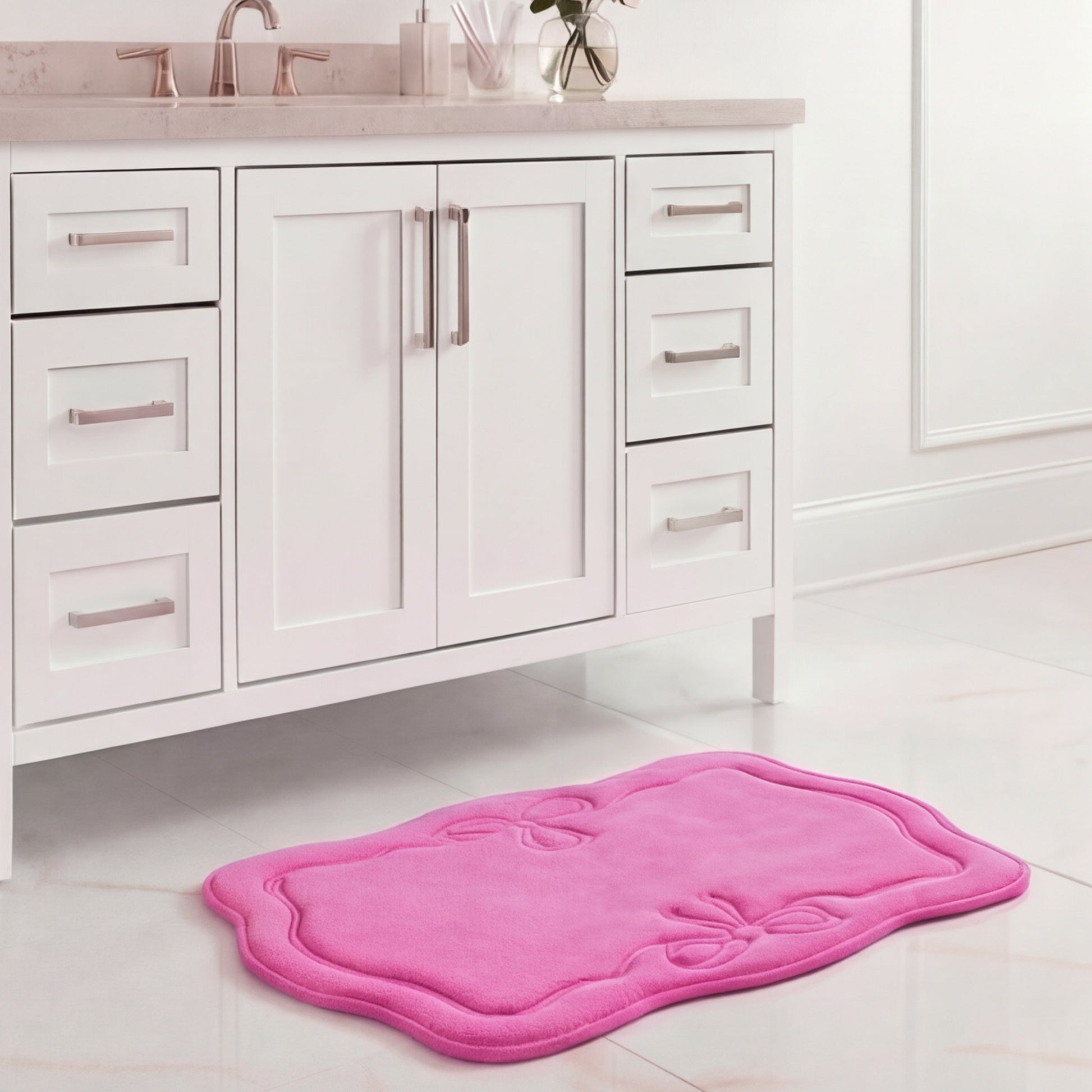 A white bathroom vanity with silver handles stands above a 17" x 24" Quilted Memory Foam Bath Mat on a white tiled floor. The pink mat's scalloped edge and embossed bow designs add luxurious style to the space.