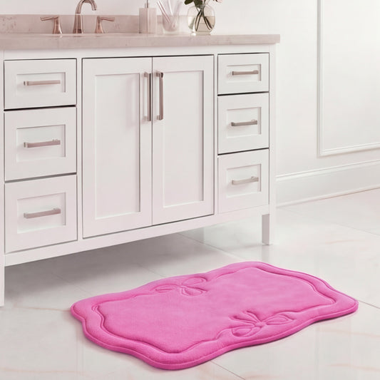 A white bathroom vanity with silver handles stands above a 17" x 24" Quilted Memory Foam Bath Mat on a white tiled floor. The pink mat's scalloped edge and embossed bow designs add luxurious style to the space.