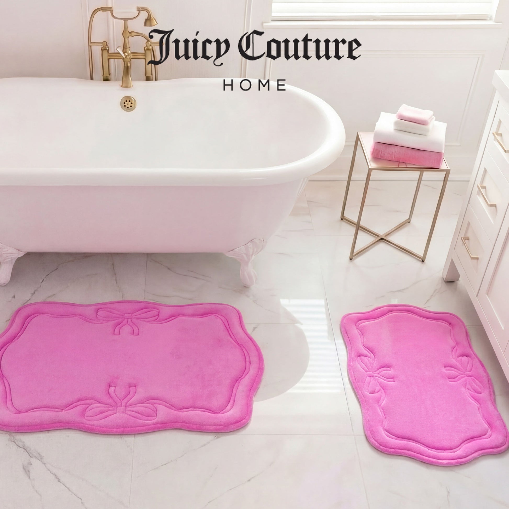 A bathroom with a white tub, gold faucet, and two Juicy Couture Quilted Memory Foam Bath Mats on a marble floor. A small table holds folded pink and white towels. “Juicy Couture Home” appears above the tub.