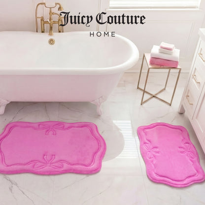 A bathroom with a white tub, gold faucet, and two Juicy Couture Quilted Memory Foam Bath Mats on a marble floor. A small table holds folded pink and white towels. “Juicy Couture Home” appears above the tub.