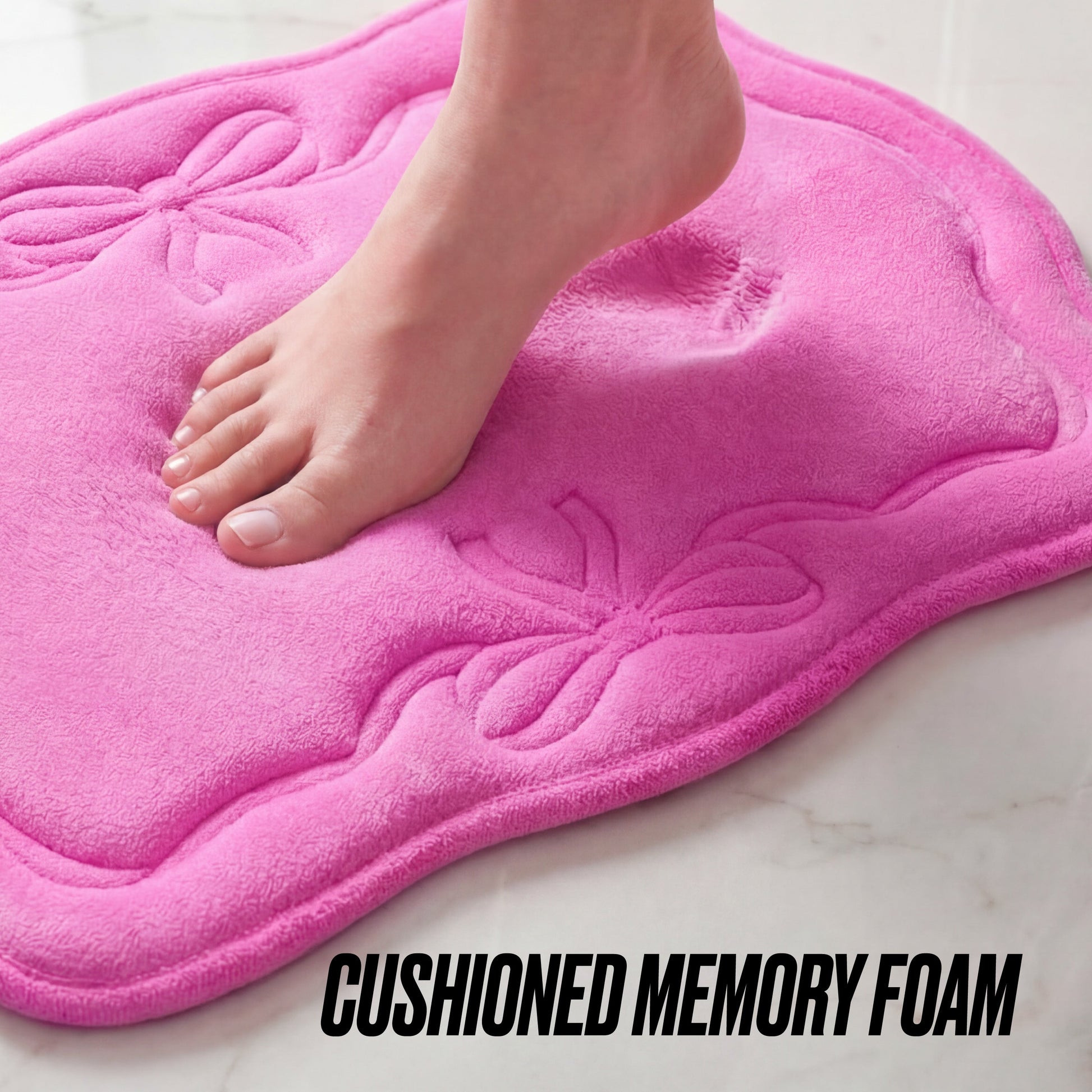 A bare foot steps on a pink Quilted Memory Foam Bath Mat 2-Piece Set, leaving an imprint in the soft, cushioned surface with embossed bows. The mat sits on a light tiled floor. Text reads: CUSHIONED MEMORY FOAM.