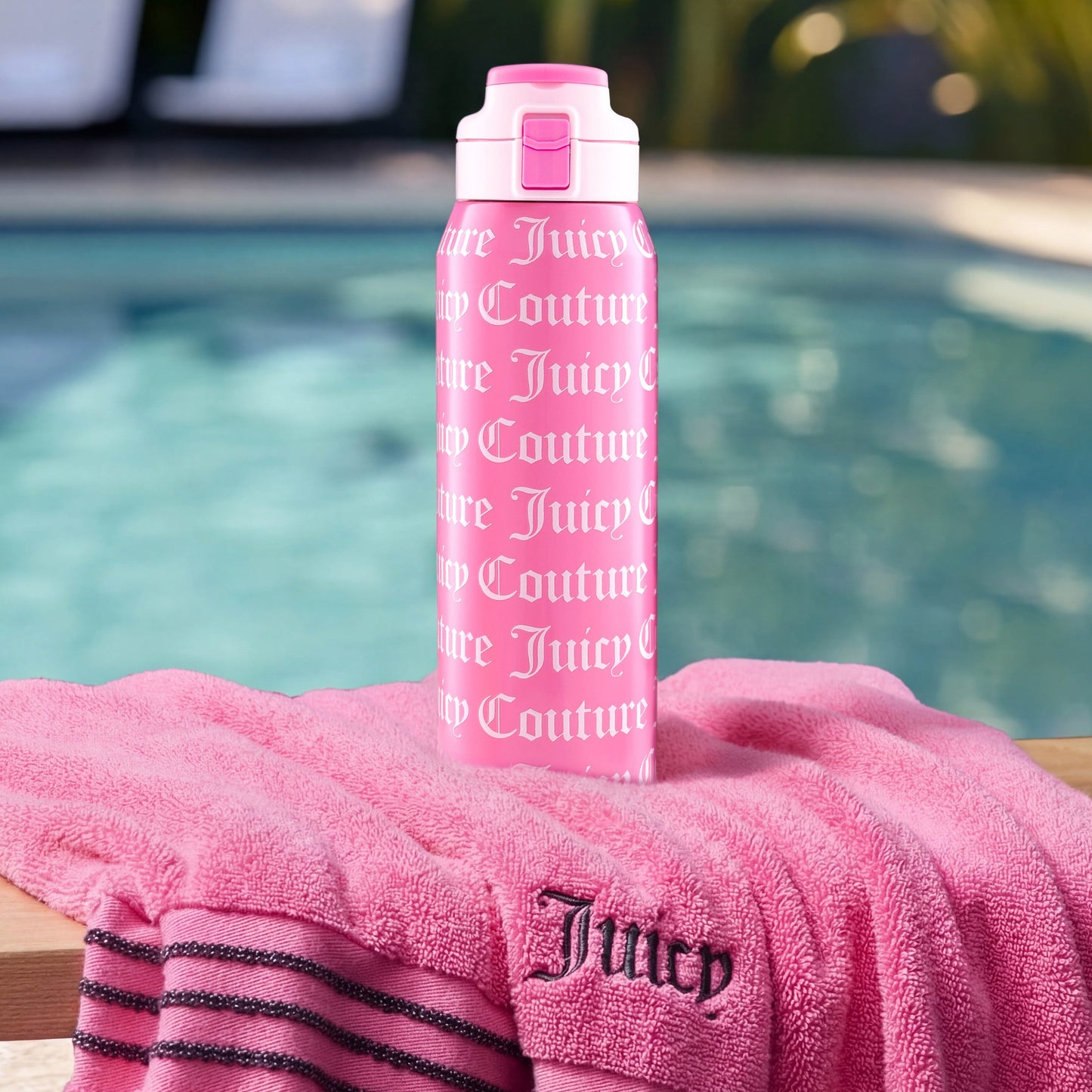 A pink Glam Getaway Hydration Stainless Steel Tumbler with white Juicy Couture logo stands on a matching pink towel draped over a wooden ledge by the pool, with lush greenery visible in the background.