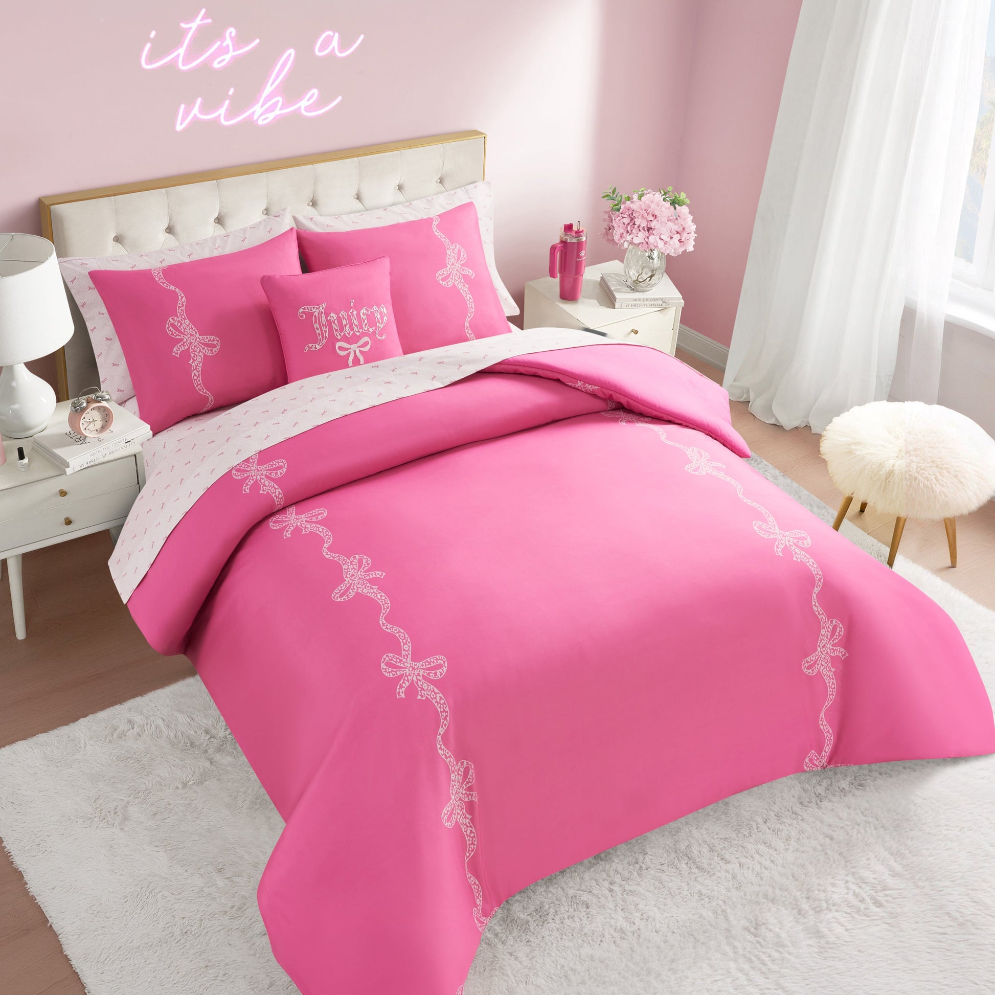 A bedroom with a bright pink Hotel Stripe Leopard Bows Bed in a Bag set, bows, white furniture, fluffy white rug, pink flowers in a vase, and a neon "its a vibe" sign above the bed. Sunlight pours through sheer white curtains.
