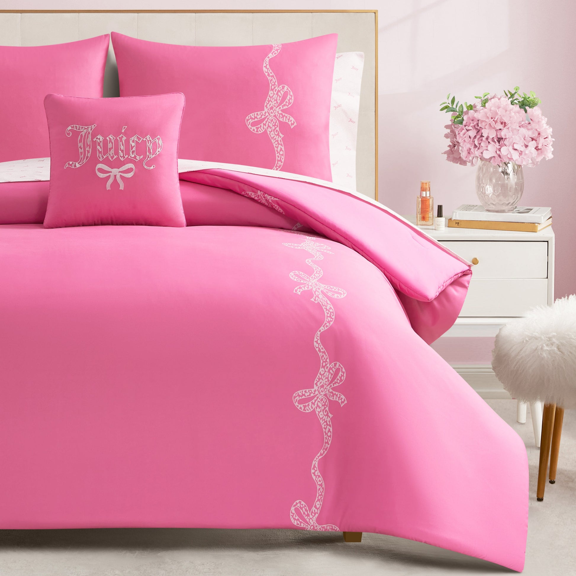 A Hotel Stripe Leopard Bows Bed in a Bag Bedding Set with white lace bow patterns and a "Juicy" pillow decorates this pink room, complete with nightstand, vase of flowers, and a white fluffy stool.