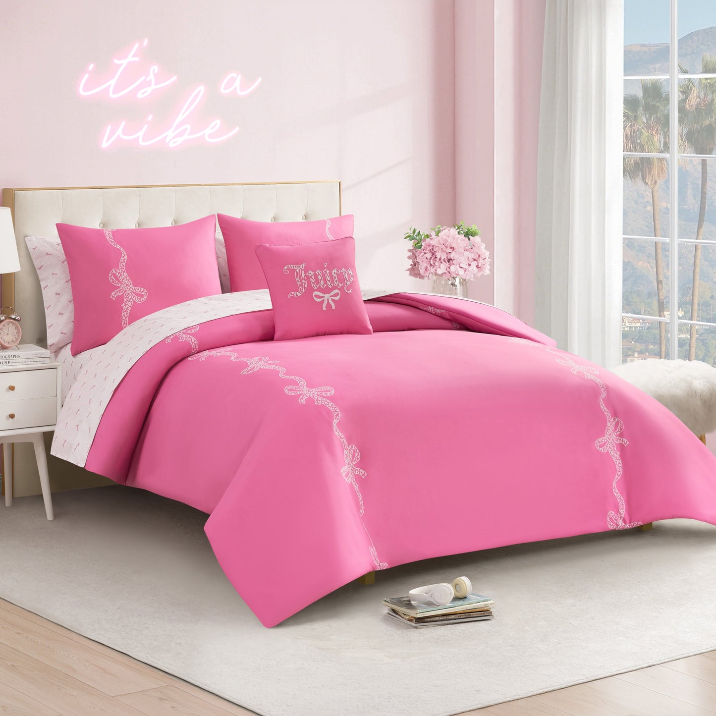 A neatly made bed with the Hotel Stripe Leopard Bows Bed in a Bag Bedding Set sits in a pastel pink room. Decorative pillows and a “it’s a vibe” neon sign are above, while sunlight streams in to reveal palm trees outside.