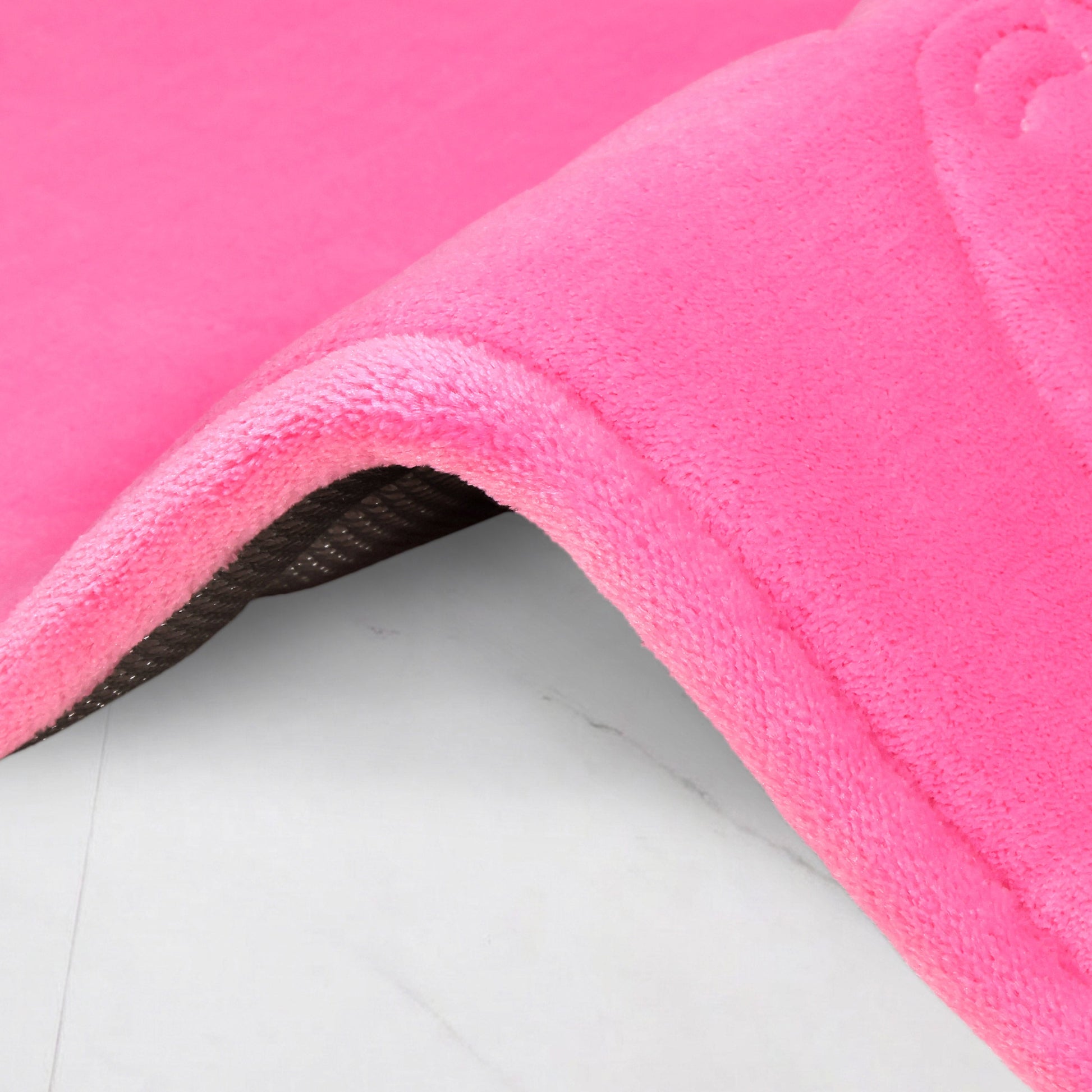 A close-up of a bright pink Quilted Memory Foam Bath Mat, 17" x 24", with a soft, plush texture and slightly curved edge, shows its dark non-slip backing as it rests on a light surface.