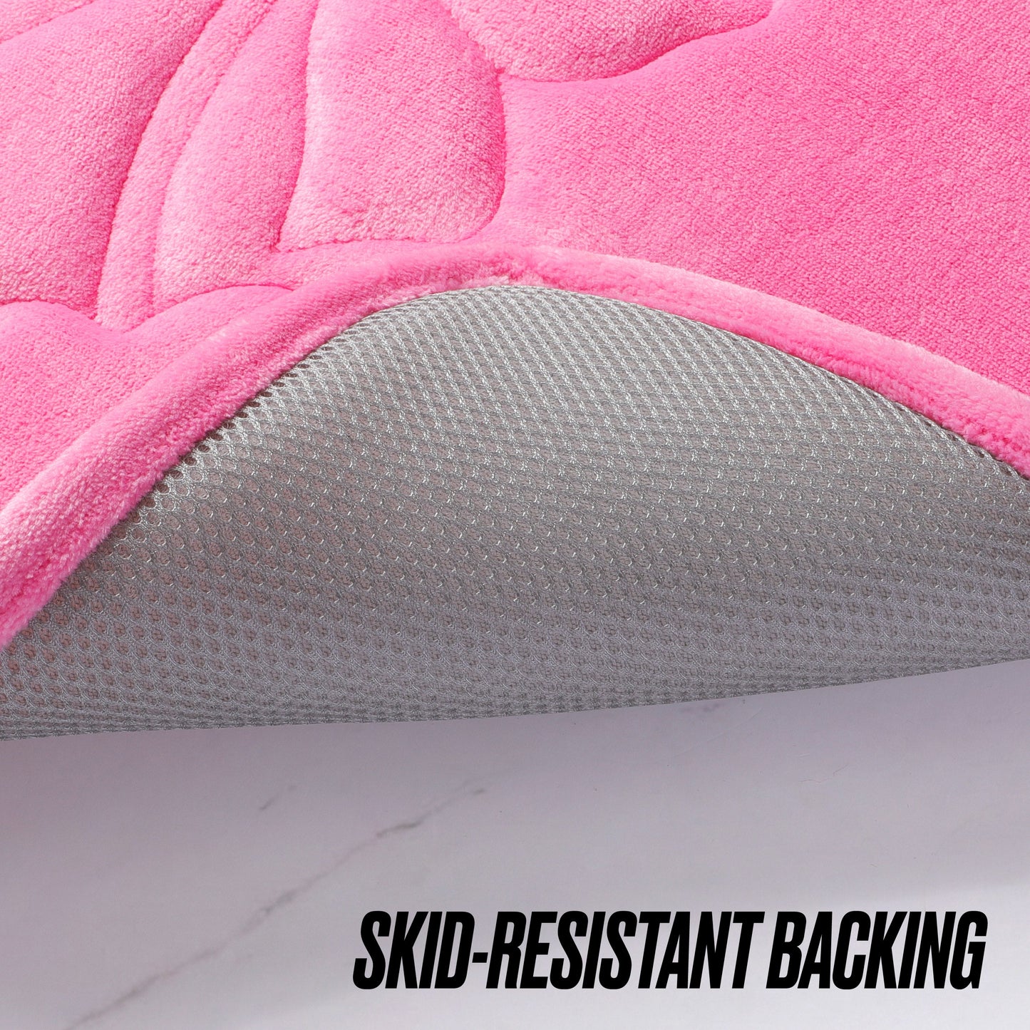 A close-up of the Quilted Memory Foam Bath Mat, 17" x 24", in pink with a corner lifted to show its textured gray skid-resistant backing. Bold black text at the bottom right reads “SKID-RESISTANT BACKING.”.