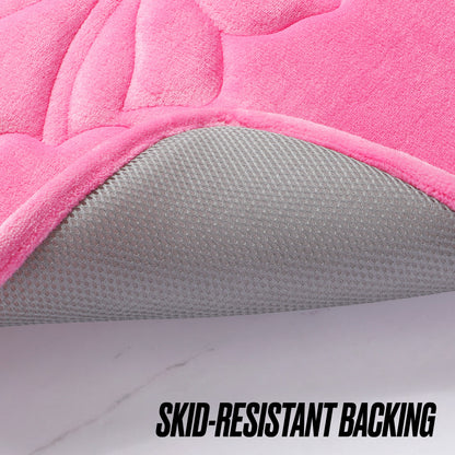 A close-up of the Quilted Memory Foam Bath Mat, 17" x 24", in pink with a corner lifted to show its textured gray skid-resistant backing. Bold black text at the bottom right reads “SKID-RESISTANT BACKING.”.
