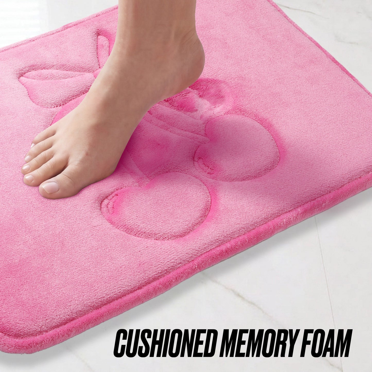A bare foot steps onto the Quilted Memory Foam Bath Mat, 17" x 24", featuring a pink quilted surface with an embossed cherry design. The cushioned mat rests on a white tile floor, with "CUSHIONED MEMORY FOAM" text at the bottom.