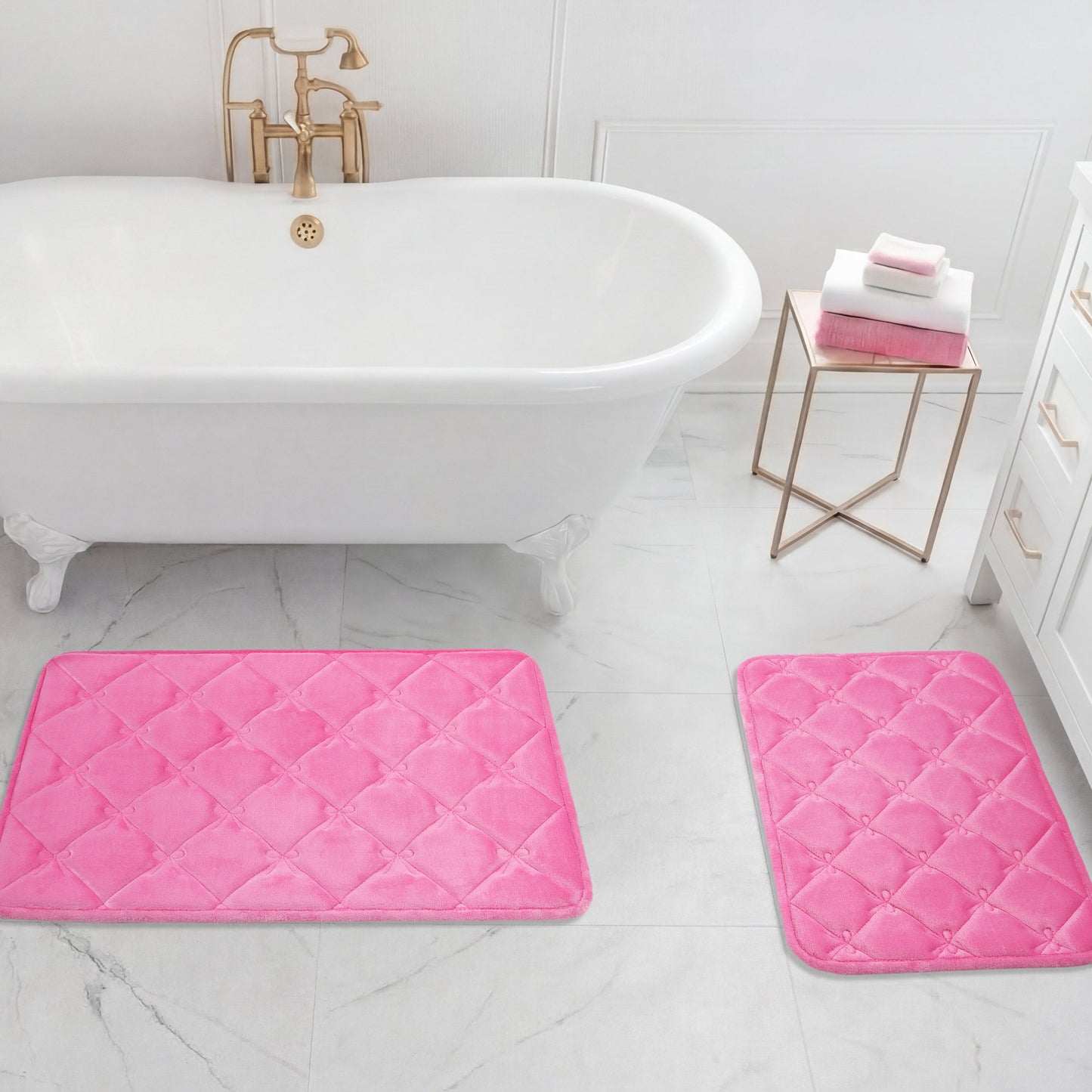 A white bathtub with gold fixtures stands next to a gold side table with folded towels. A Quilted Memory Foam Bath Mat 2-Piece Set in pink is placed on the white tile floor in this bright bathroom.