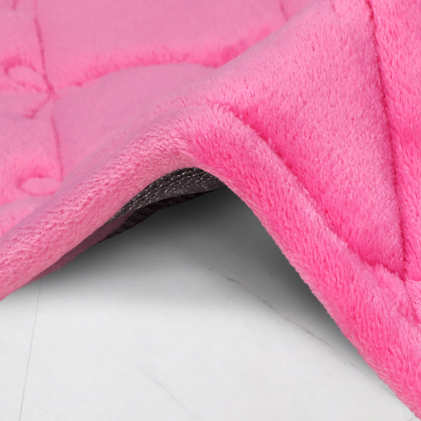 Close-up of the Quilted Memory Foam Bath Mat 2-Piece Set in soft pink, featuring plush quilting and a raised edge with textured gray underside, shown on a light-colored surface.