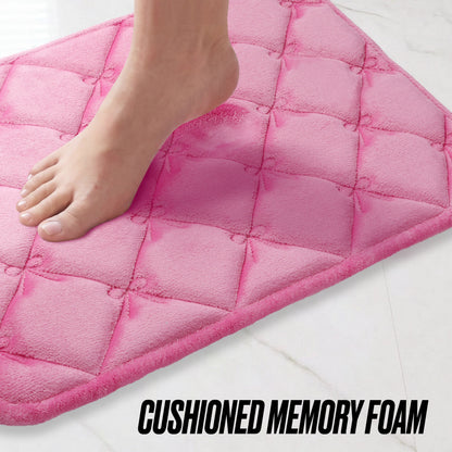 A bare foot steps onto a pink Quilted Memory Foam Bath Mat 2-Piece Set on a white tiled floor. The text at the bottom reads CUSHIONED MEMORY FOAM.