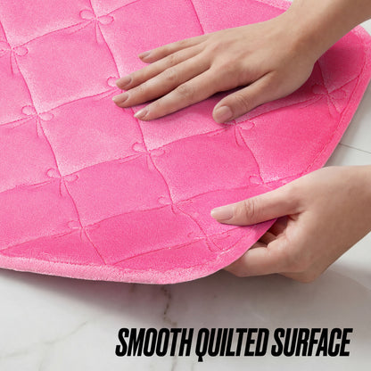 A person touches the corner of a Quilted Memory Foam Bath Mat 2-Piece Set in bright pink, highlighting its smooth, quilted surface. "SMOOTH QUILTED SURFACE" appears in bold black text at the bottom.