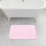 A pink Quilted Memory Foam Bath Mat, 17" x 24", with six embossed bow designs is placed on a light gray marble floor in front of a white clawfoot bathtub.