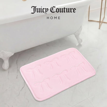 A pale pink Juicy Couture Quilted Memory Foam Bath Mat, 17" x 24", with embossed bow designs rests on a white marble floor beside a white clawfoot tub. "Juicy Couture HOME" is displayed above the bathtub.