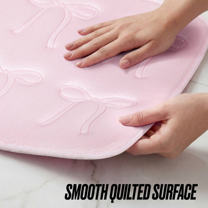 Two hands gently touch a light pink Quilted Memory Foam Bath Mat, 17" x 24", featuring an embossed bow pattern and a soft, quilted surface. The text "SMOOTH QUILTED SURFACE" appears at the bottom of the image.