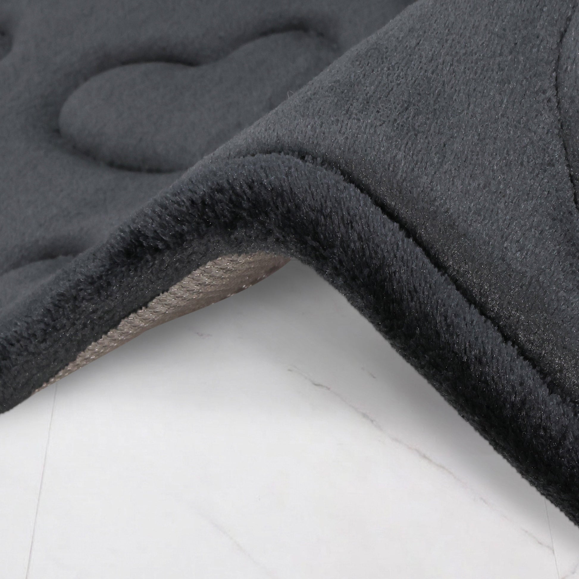 Close-up of the Quilted Memory Foam Bath Mat 2-Piece Set in dark gray, featuring thick, plush memory foam with a quilted texture and a slightly raised edge, shown lying on a light surface. The mats look soft and cushioned.