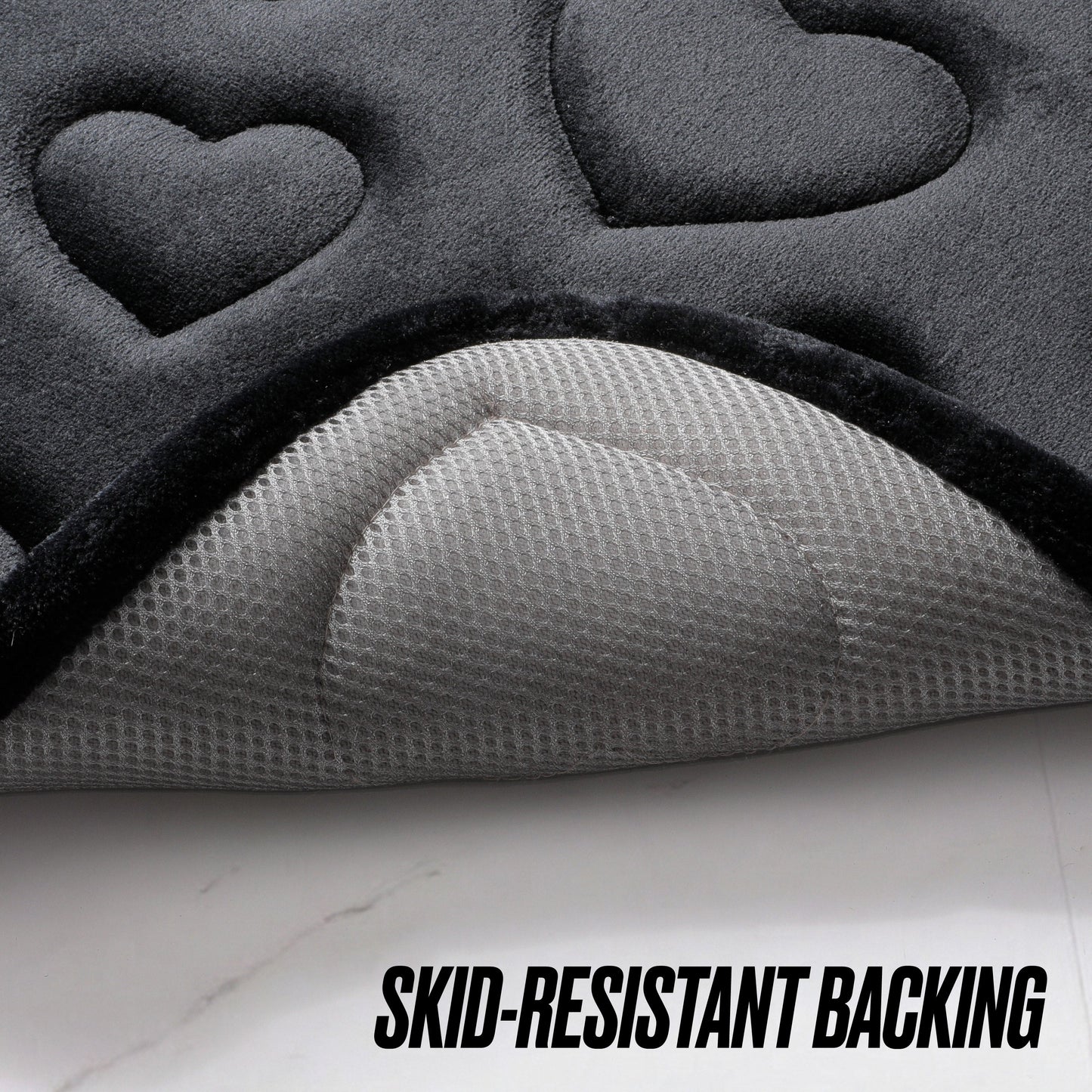 Close-up of the Quilted Memory Foam Bath Mat 2-Piece Set in a dark color with embossed heart shapes, partially lifted to reveal its textured, light gray skid-resistant backing. Text reads: SKID-RESISTANT BACKING.
