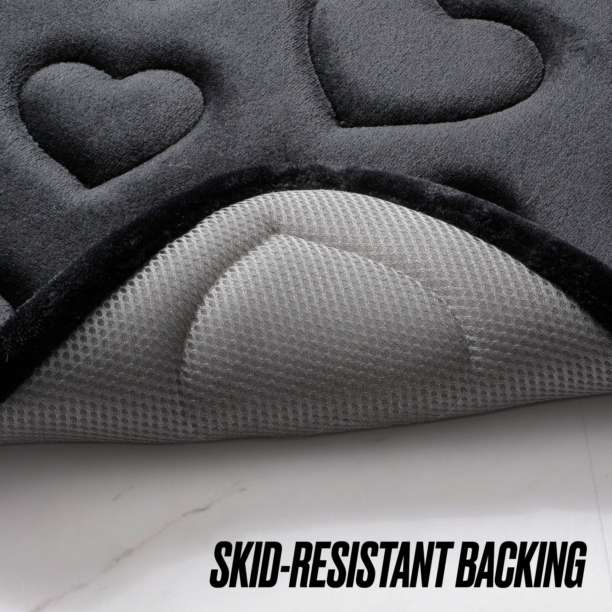 Close-up of the Quilted Memory Foam Bath Mat 2-Piece Set in a dark color with embossed heart shapes, partially lifted to reveal its textured, light gray skid-resistant backing. Text reads: SKID-RESISTANT BACKING.