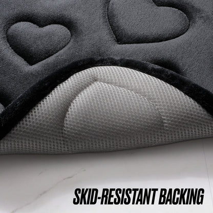 Close-up of the Quilted Memory Foam Bath Mat 2-Piece Set in a dark color with embossed heart shapes, partially lifted to reveal its textured, light gray skid-resistant backing. Text reads: SKID-RESISTANT BACKING.