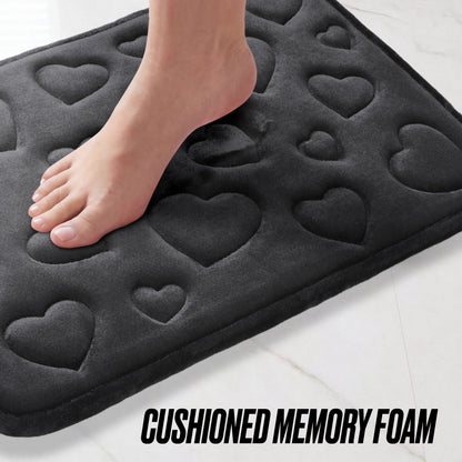A bare foot steps onto the Quilted Memory Foam Bath Mat 2-Piece Set, featuring soft black quilted memory foam with embossed heart shapes. The text "CUSHIONED MEMORY FOAM" appears at the bottom of the image.