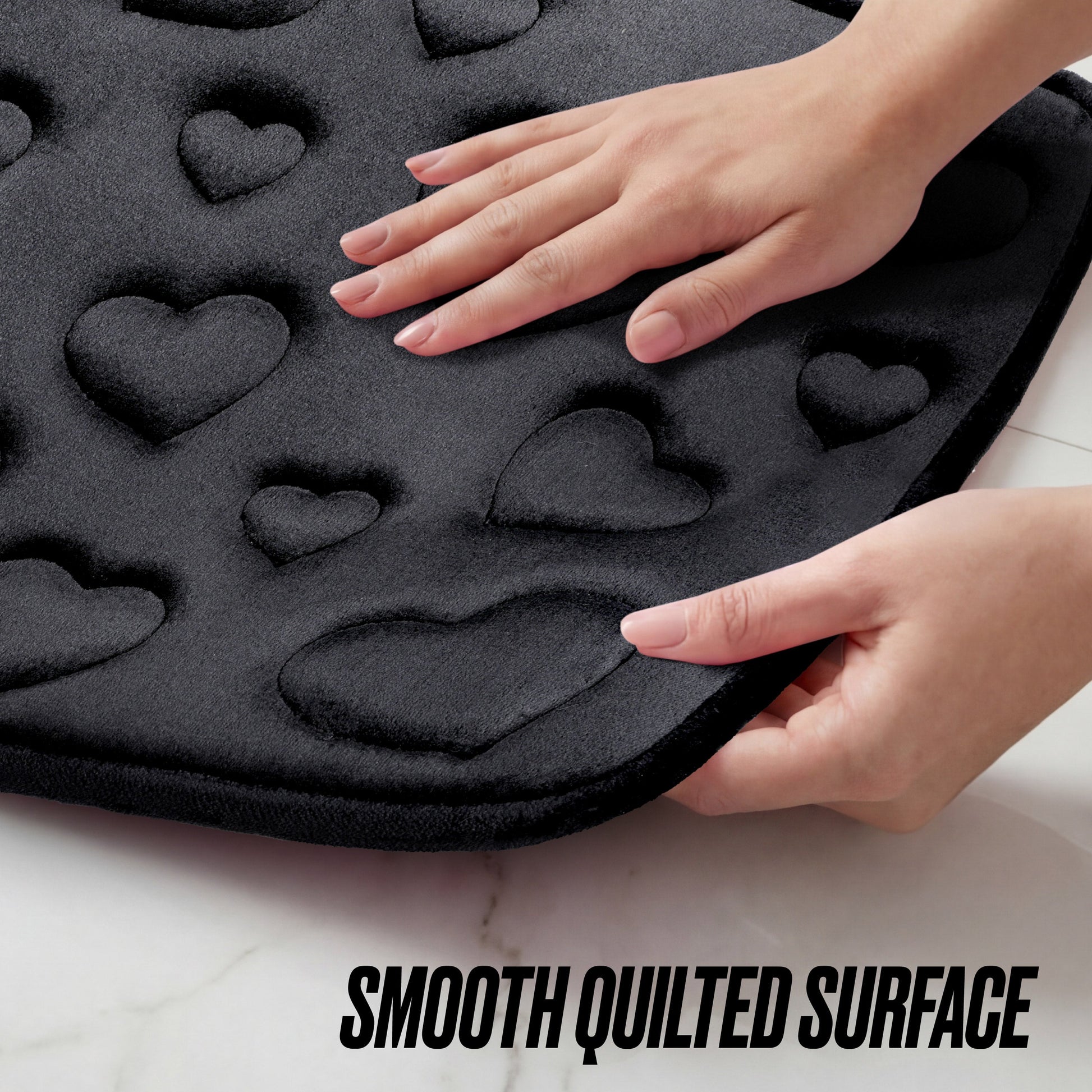 Two hands touch a soft, black Quilted Memory Foam Bath Mat 2-Piece Set featuring a quilted pattern of raised hearts on a white surface. Text at the bottom reads “Smooth Quilted Surface.”.