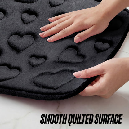 Two hands touch a soft, black Quilted Memory Foam Bath Mat 2-Piece Set featuring a quilted pattern of raised hearts on a white surface. Text at the bottom reads “Smooth Quilted Surface.”.