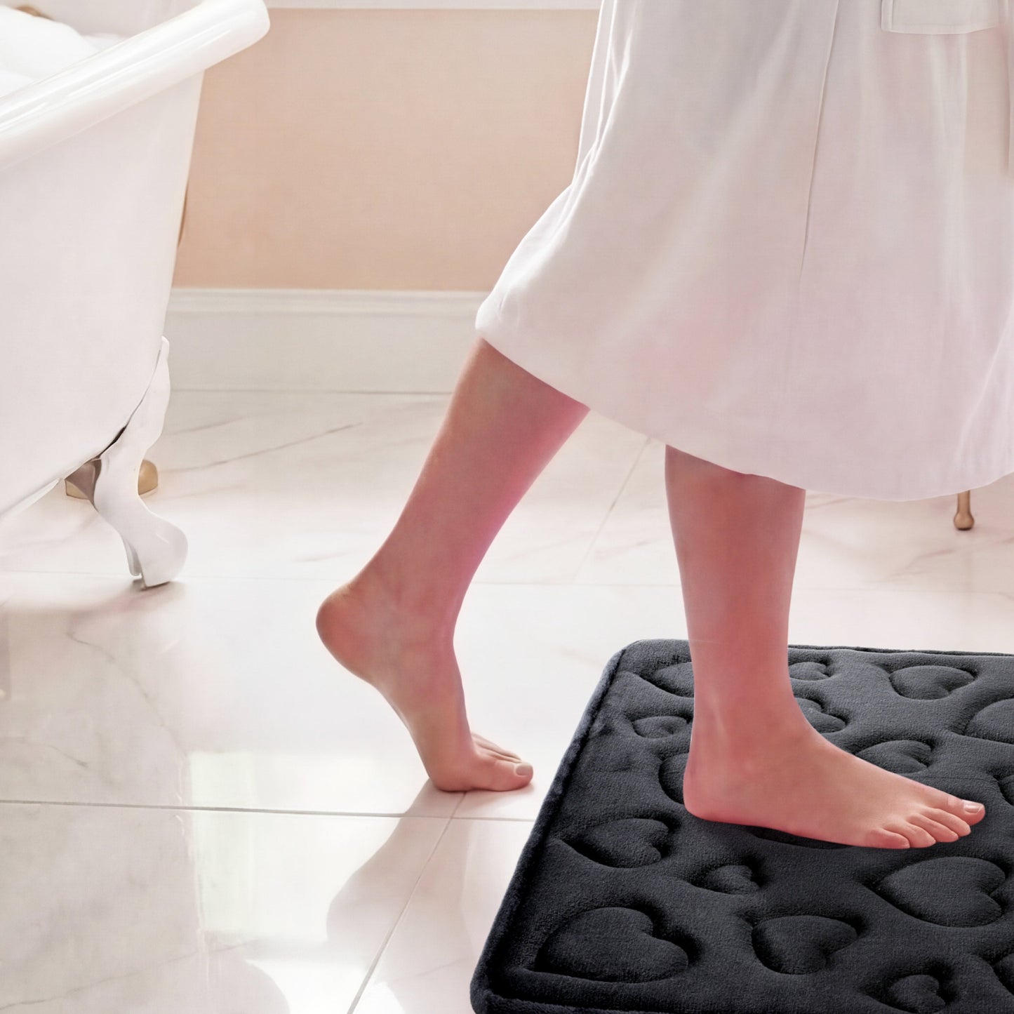 A person in a white robe steps onto the Quilted Memory Foam Bath Mat 2-Piece Set with bare feet in a bright, clean bathroom featuring a bathtub and tiled floor.