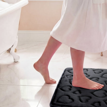 A person in a white robe steps onto the Quilted Memory Foam Bath Mat 2-Piece Set with bare feet in a bright, clean bathroom featuring a bathtub and tiled floor.