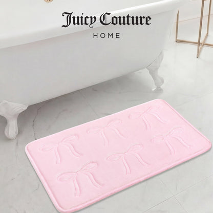 A Quilted Memory Foam Bath Mat (20" x 32") in light pink, featuring embossed bows, rests on a marble bathroom floor near a white clawfoot tub. "Juicy Couture Home" is elegantly displayed above the mat.