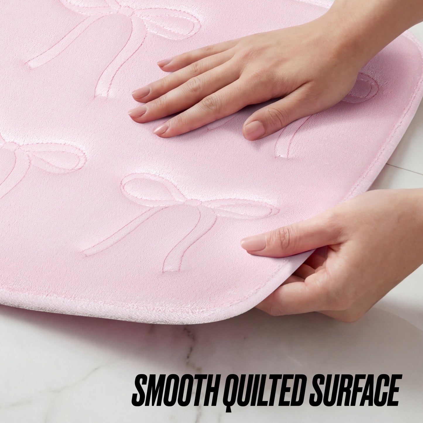 Hands touch the light pink Quilted Memory Foam Bath Mat, 20" x 32", featuring a stylish quilted bow pattern on a white surface. The text SMOOTH QUILTED SURFACE appears at the bottom.