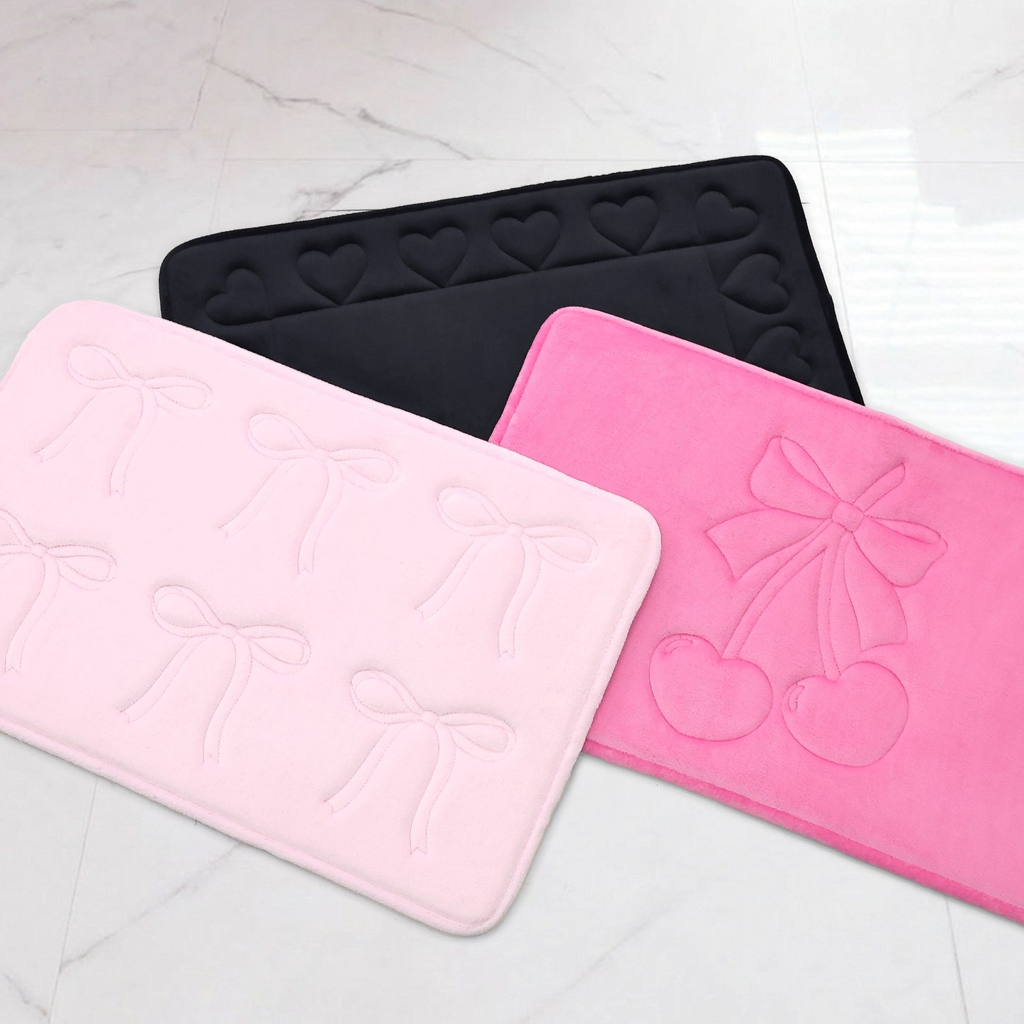A black Juicy Couture bath mat with hearts, a light pink plush rug with bows, and a hot pink mat with a bow and cherries are shown on a white floor; the light pink one is the Quilted Memory Foam Bath Mat, 20" x 32".