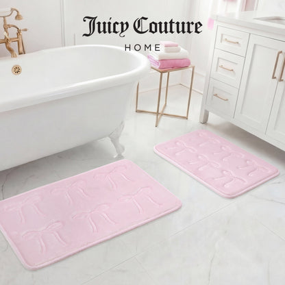 A white bathroom features a clawfoot tub, white vanity, and two light pink Juicy Couture Quilted Memory Foam Bath Mats. Folded pink and white towels rest on the vanity. “Juicy Couture Home” is written at the top.