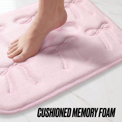 A bare foot steps onto a soft, light pink Quilted Memory Foam Bath Mat 2-Piece Set with bow patterns, placed on a white tiled floor. The text CUSHIONED MEMORY FOAM appears at the bottom.