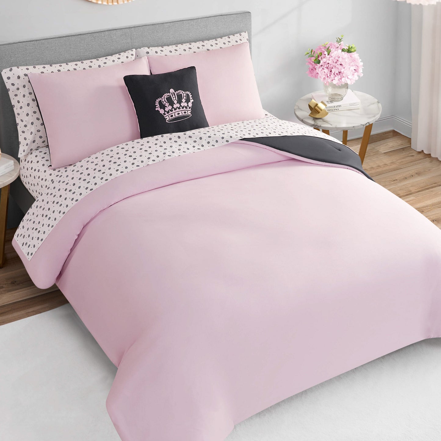 A neatly made bed with the Crown Sparkle Solid Reversible Bed in a Bag Bedding Set features a light pink comforter, matching pillows, a decorative black crown pillow, and white sheets with black hearts. Flowers and natural light add glam to the space.