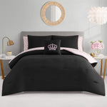 A neatly made Crown Sparkle Solid Reversible Bed in a Bag Bedding Set features black bedding, a pink and black polka dot sheet, and a crown pillow. Glam decor includes a gray headboard, gold accents, round mirror, and pink nightstand flowers.