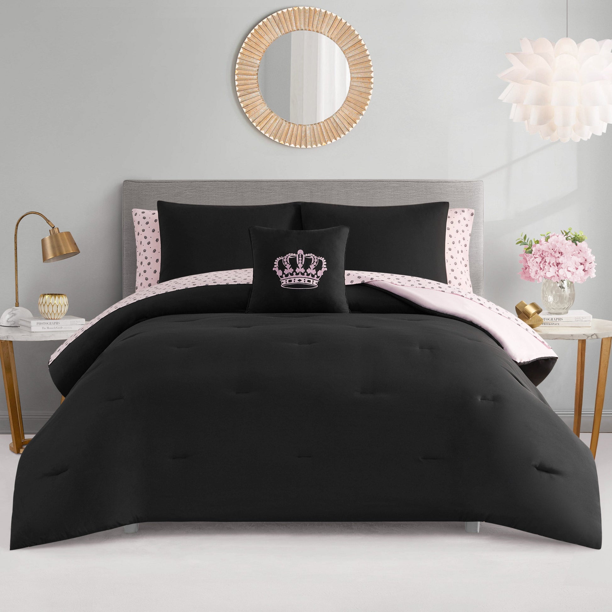 A neatly made Crown Sparkle Solid Reversible Bed in a Bag Bedding Set features black bedding, a pink and black polka dot sheet, and a crown pillow. Glam decor includes a gray headboard, gold accents, round mirror, and pink nightstand flowers.
