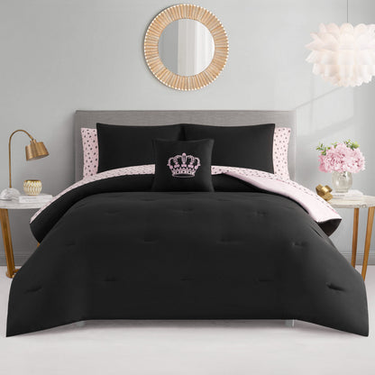 A neatly made Crown Sparkle Solid Reversible Bed in a Bag Bedding Set features black bedding, a pink and black polka dot sheet, and a crown pillow. Glam decor includes a gray headboard, gold accents, round mirror, and pink nightstand flowers.