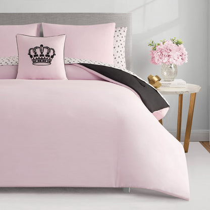 A neatly made bed features the Crown Sparkle Solid Reversible Bed in a Bag Bedding Set with light pink bedding, a pillow with a black crown design, and a polka dot reversible duvet. A nightstand with pink flowers and books sits to the right.